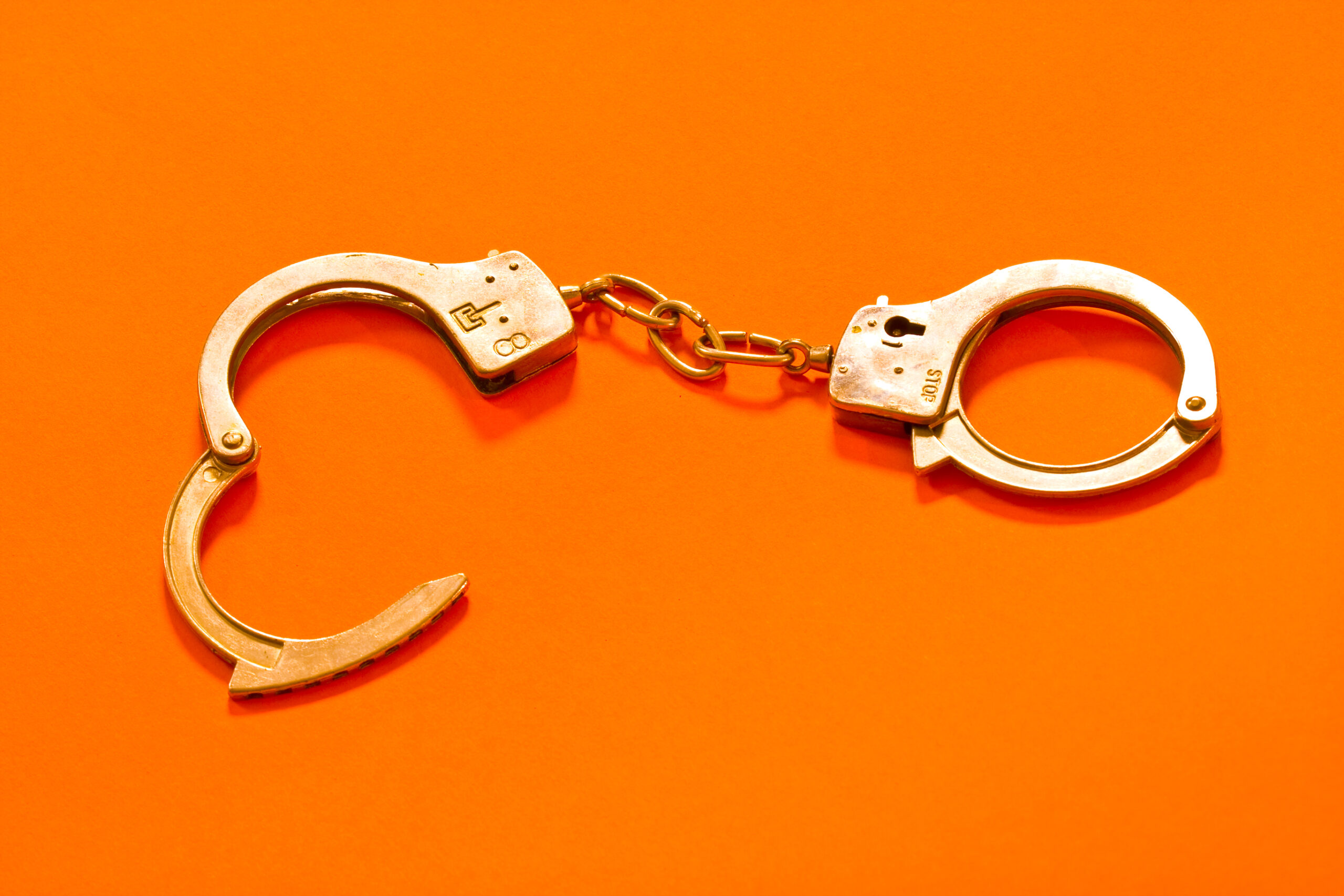 Two metal handcuffs connected by a short chain on a bright orange background; one cuff is open.